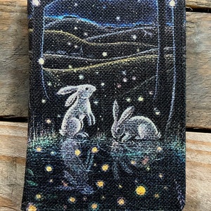 May include: A black canvas journal with a whimsical illustration of two white rabbits in a field of glowing fireflies under a starry night sky.
