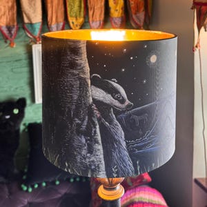 May include: A lampshade with a badger illustration against a night sky backdrop. The lampshade has a gold interior and is mounted on a wooden base. The design creates a nature-inspired aesthetic, suitable for a living room or bedroom.