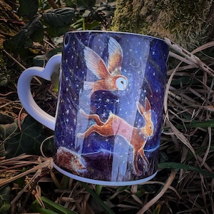 Könnte beinhalten: Weiße Keramiktasse mit einem herzförmigen Henkel. Die Tasse zeigt eine farbenfrohe Illustration eines fliegenden Eulen, eines laufenden Hasen und eines Igels vor einem dunkelblauen Hintergrund mit weißen Sternen.