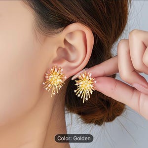 Puede incluir: Pendientes dorados con diseño de estrella. Los pendientes presentan un grupo central de pequeños cristales transparentes rodeados de puntas doradas radiantes. El texto "Color: Golden" es visible.