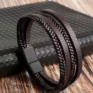May include: A multi-strand brown leather bracelet with a magnetic clasp. The bracelet features braided and smooth leather strands. The clasp is black. The bracelet is shown on a textured black surface.