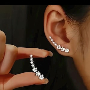 May include: A silver-toned ear crawler earring featuring a curved row of five clear, sparkling, flower-shaped crystals. The earring follows the natural curve of the earlobe, with each flower set with a central stone surrounded by petals.