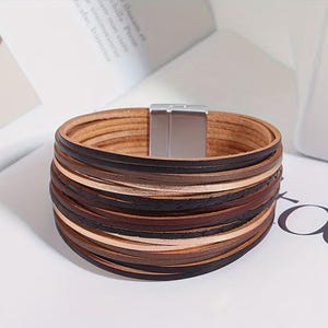 May include: A multi-layered leather bracelet with a magnetic clasp. The bracelet features various shades of brown leather strips, including light tan, dark brown, and black. The clasp is silver-colored. The bracelet is a stylish accessory.