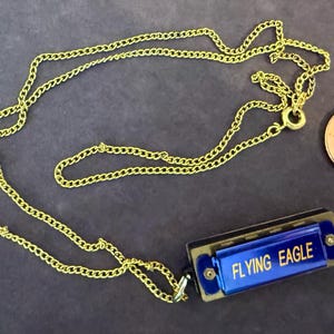 May include: A gold-colored chain necklace with a small blue harmonica pendant. The harmonica has the words "FLYING EAGLE" in white text. A penny is next to the necklace for size comparison.