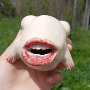 May include: A whimsical, off-white ceramic sculpture of a creature with a wide open mouth and red lipstick. The sculpture has small arms and feet. The piece is held in a hand against a blurred outdoor background.