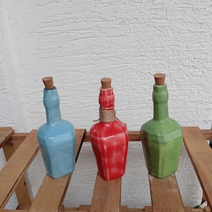 May include: Three decorative ceramic bottles in blue, red, and green, each with a cork stopper. The bottles have unique geometric shapes and are displayed on a wooden surface. The red bottle has twine wrapped around its neck.