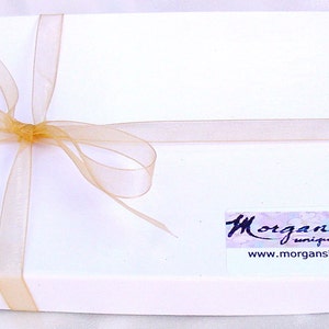 以下が含まれることがあります： 金色のリボンが結ばれた白いギフトボックス。ボックスには「Morgan Silk unique scarves」と「www.morgansilkscarf.com」と書かれたラベルが付いています。