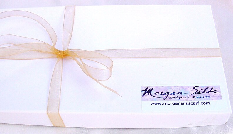 以下が含まれることがあります： 金色のリボンが結ばれた白いギフトボックス。ボックスには「Morgan Silk unique scarves」と「www.morgansilkscarf.com」と書かれたラベルが付いています。