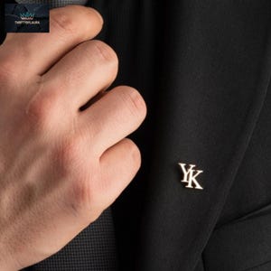 Może przedstawiać: Srebrna przypinka do klapy z inicjałami "YK" na czarnej marynarce. Przypinka jest mała i elegancka, dodając odrobinę wyrafinowania formalnemu strojowi. Marynarka jest częścią garnituru.