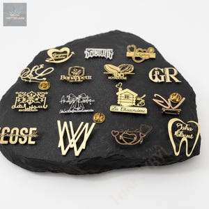 May include: A selection of gold and silver-coloured metal pins with diverse designs, including text and symbols, presented on a black, textured surface. The pins feature words like "Bonappettt" and "Dr Galber."