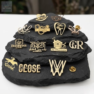 May include: A collection of gold-colored pins and brooches displayed on a black, textured surface. The pins feature various designs, including medical symbols, text, and decorative shapes. Some pins have text like "Dr Gallier" and "Bonappetit".