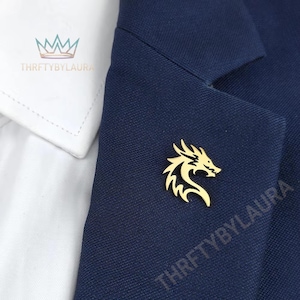 May include: A gold-colored dragon lapel pin on a navy blue blazer. The pin is a stylized depiction of a dragon with intricate details. The blazer is paired with a white collared shirt. The shop name "THRIFTYBYLAURA" is visible.