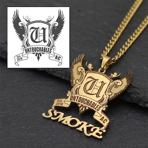 Op de afbeelding: Goudkleurige hangerketting met een gedetailleerd ontwerp. De hanger heeft een schild met het woord "UNTOUCHABLES" en het woord "SMOKE" in een gestileerd lettertype. De ketting bevat een ketting en een logo in de linkerbovenhoek.