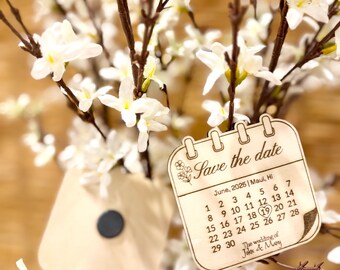 May include: Wooden save the date card with a calendar design. The card is engraved with the text "Save the date" and the date "June, 2025 | Maui, Hi". The calendar shows the date "19" circled. The text "The wedding of John & Mary" is also engraved on the card.