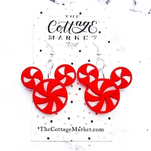 May include: Red and white candy cane shaped earrings with silver hooks. The earrings are shaped like Mickey Mouse ears. The earrings are on a white card with the text "The Cottage Market" and "TheCottageMarket.com".