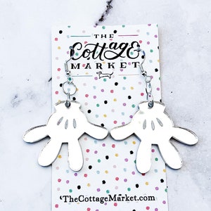 May include: Silver earrings with a white, mirrored Mickey Mouse hand design. The earrings are on a card with the text "The Cottage Market" and a website address.