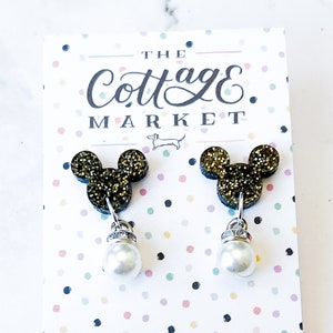 Może przedstawiać: Para złotych kolczyk&oacute;w z motywem Myszki Miki z perłową zawieszką. Kolczyki znajdują się na białej kartce z tekstem "The Cottage Market" i "TheCottageMarket.com".