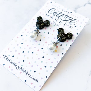 Może przedstawiać: Czarne błyszczące kolczyki z motywem Myszki Miki z perłowym wisiorem. Kolczyki znajdują się na białej kartce z nadrukiem "The Cottage Market".