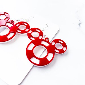 May include: Red and white Mickey Mouse-shaped earrings. The earrings feature a circular design with a white center and red outer ring. The earrings are made of a shiny material and have silver-colored hooks.