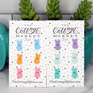May include: A set of six pastel-colored bunny-shaped earrings. The earrings are displayed on a white card with the text "THE Cottage MARKET" and "TheCottageMarket.com".