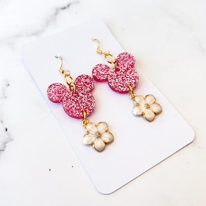 Puede incluir: Pendientes de Mickey Mouse con purpurina rosa y abalorios de flores blancas. Los pendientes est&aacute;n sobre un fondo blanco.