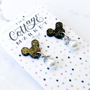 Może przedstawiać: Para złotych kolczyk&oacute;w z motywem Myszki Miki z perłowymi akcentami. Kolczyki są prezentowane na kartce z tekstem "The Cottage Market".