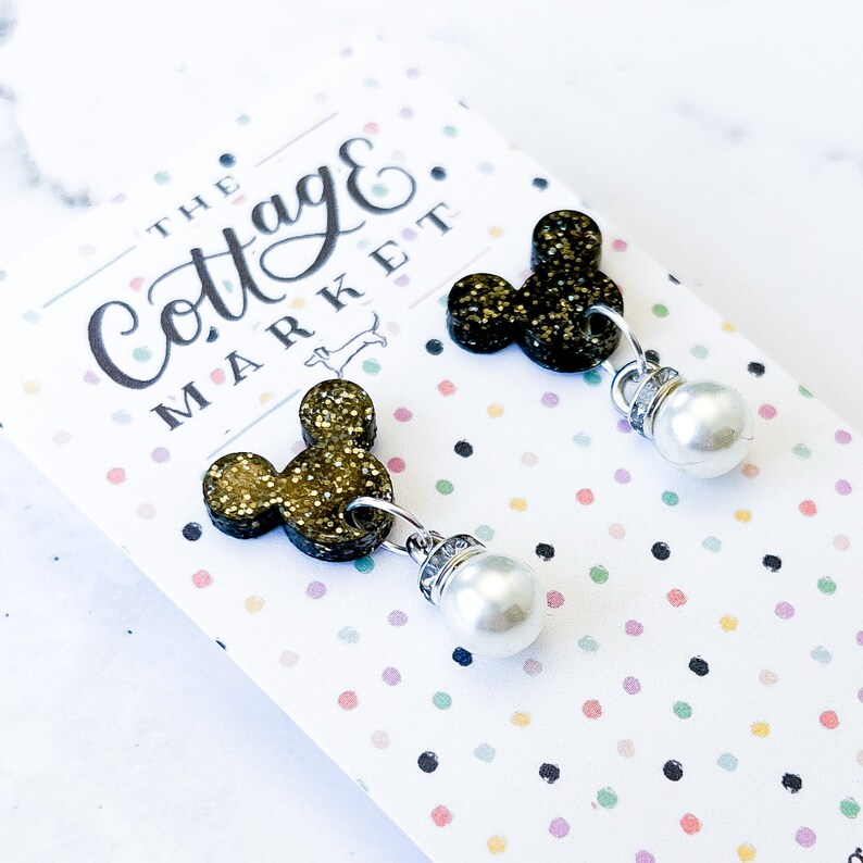 Może przedstawiać: Para złotych kolczyk&oacute;w z motywem Myszki Miki z perłowymi akcentami. Kolczyki są prezentowane na kartce z tekstem "The Cottage Market".