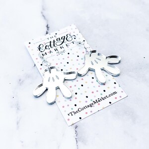 May include: Silver mirror acrylic earrings shaped like Mickey Mouse's iconic white gloves. The earrings are hanging from a card with the text "The Cottage Market".