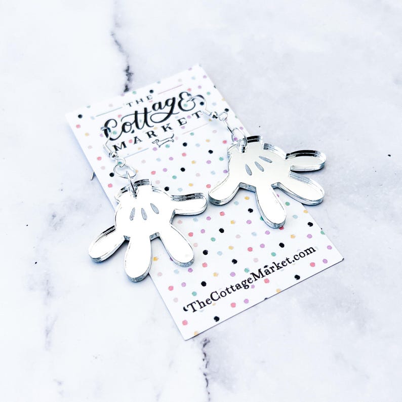 May include: Silver mirror acrylic earrings shaped like Mickey Mouse's iconic white gloves. The earrings are hanging from a card with the text "The Cottage Market".