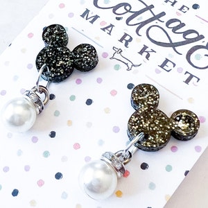 Może przedstawiać: Para czarnych błyszczących kolczyk&oacute;w z motywem Myszki Miki z perłowymi elementami. Kolczyki znajdują się na białej kartce z napisem "The Cottage Market" w kolorze czarnym.