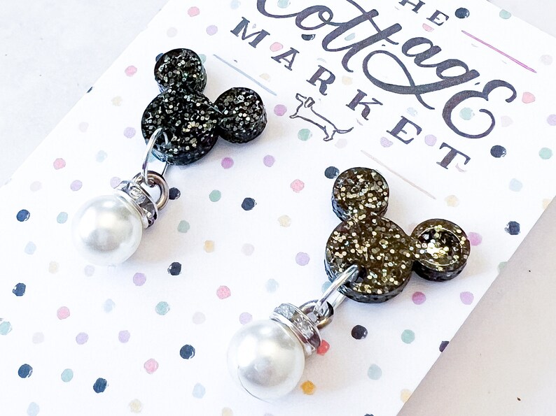 Może przedstawiać: Para czarnych błyszczących kolczyk&oacute;w z motywem Myszki Miki z perłowymi elementami. Kolczyki znajdują się na białej kartce z napisem "The Cottage Market" w kolorze czarnym.