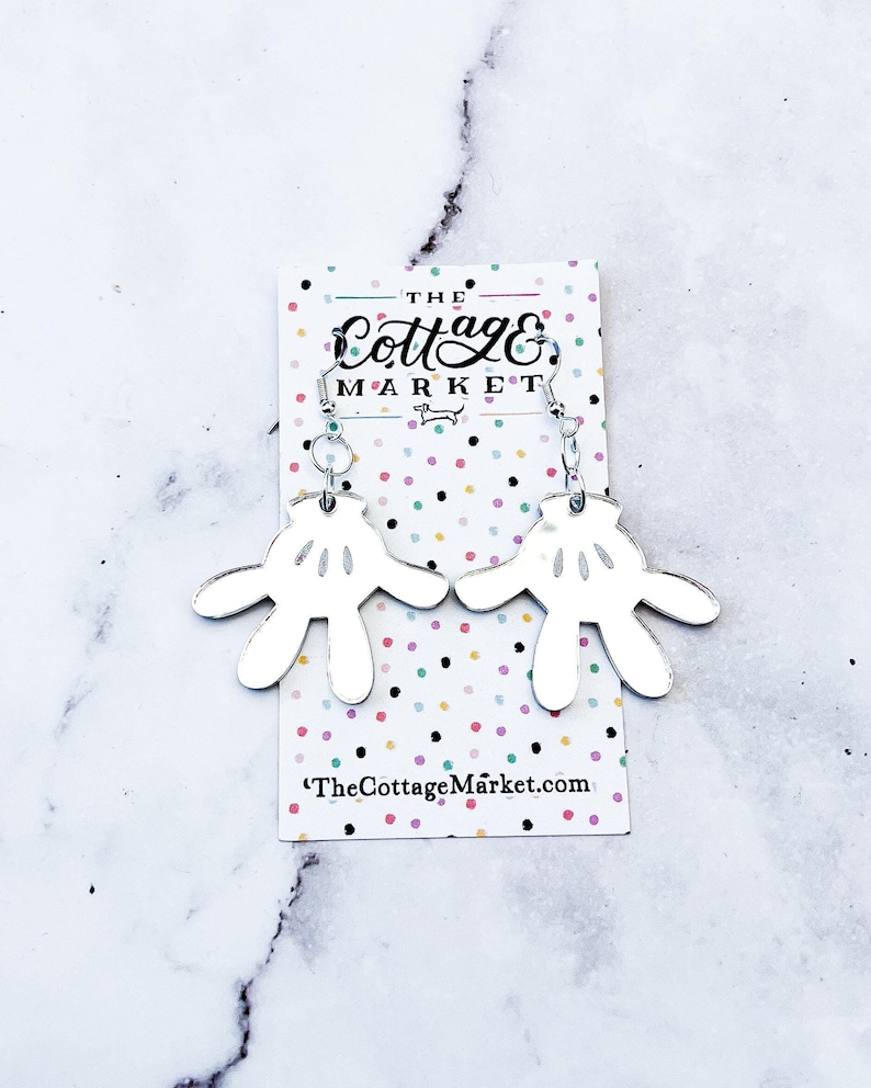 May include: Silver earrings with a white, mirrored Mickey Mouse hand design. The earrings are on a card with the text "The Cottage Market" and a website address.
