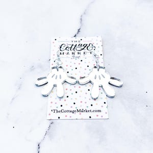 May include: Silver mirror acrylic earrings shaped like Mickey Mouse's iconic white gloves. The earrings are hanging from silver chains and have a small silver charm at the top. The earrings are displayed on a card with the text "The Cottage Market" and the website address "TheCottageMarket.com".