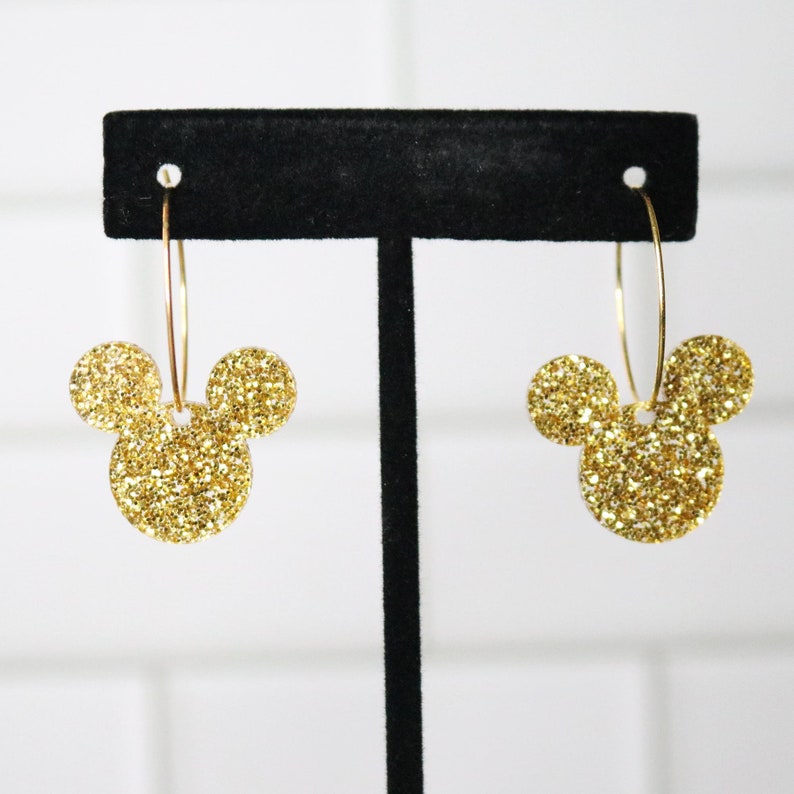 May include: Gold glitter Mickey Mouse head hoop earrings. The earrings are on a black jewelry display stand.