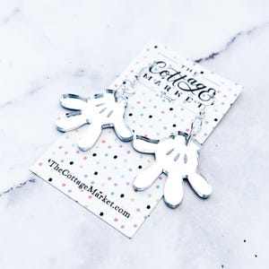 May include: Silver mirror acrylic earrings shaped like Mickey Mouse's iconic white gloves. The earrings are hanging from silver hooks. The earrings are on a white background with colorful polka dots. The text "The Cottage Market.com" is printed on the background.