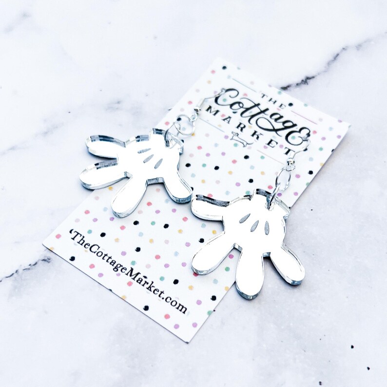 May include: Silver mirror acrylic earrings shaped like Mickey Mouse's iconic white gloves. The earrings are hanging from silver hooks. The earrings are on a white background with colorful polka dots. The text "The Cottage Market.com" is printed on the background.