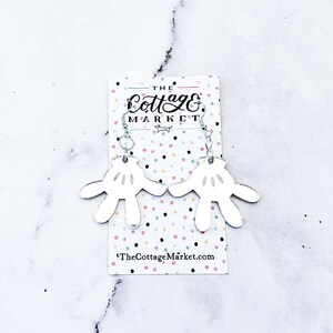 May include: Silver earrings with a white, mirrored, hand-shaped design. The earrings are on a card with the text "The Cottage Market" and a website address.