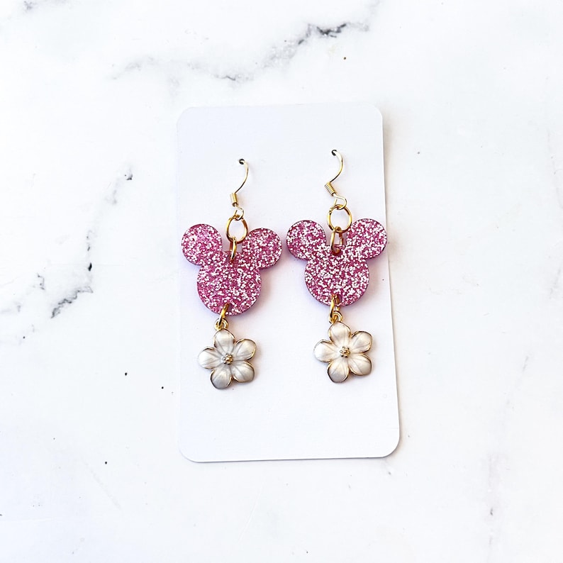 Puede incluir: Pendientes de Mickey Mouse de purpurina rosa con abalorios de flores blancas que cuelgan de ganchos dorados.