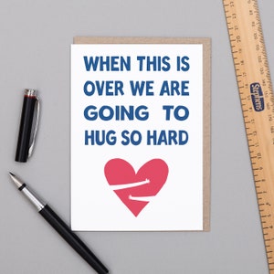 Puede incluir: Una tarjeta de felicitación con el texto "WHEN THIS IS OVER WE ARE GOING TO HUG SO HARD" en azul. Un corazón rojo con brazos abrazando está debajo del texto. Un bolígrafo negro y una regla están sobre la superficie.