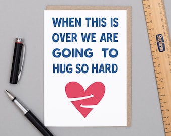 May include: A greeting card with the text "WHEN THIS IS OVER WE ARE GOING TO HUG SO HARD" in blue. A red heart with arms hugging is below the text. A black pen and a ruler are on the surface.