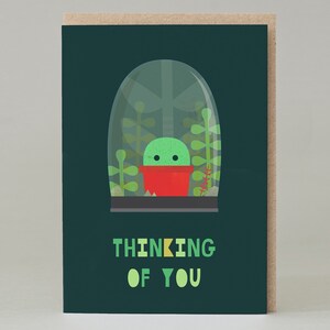 Puede incluir: Tarjeta de felicitación con un cactus de dibujos animados en una maceta roja bajo una cúpula de cristal, sobre un fondo verde oscuro. Las palabras "THINKING OF YOU" están impresas en verde y amarillo.