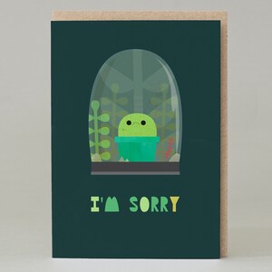 Puede incluir: Una tarjeta de felicitación con un fondo verde oscuro y un cactus de dibujos animados dentro de un terrario de cristal. El texto "I'M SORRY" está impreso en verde y amarillo. La tarjeta tiene un borde marrón.