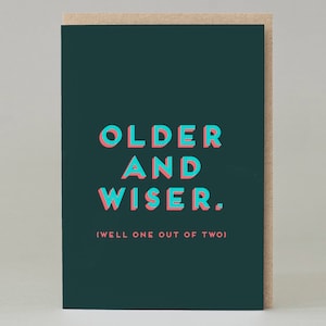 May include: Greeting card with a dark teal background and the words "OLDER AND WISER" in large, 3D aqua and pink letters. Below, in smaller text, it reads "(WELL ONE OUT OF TWO)". The card is set against a light brown background.