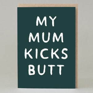 Puede incluir: Tarjeta de felicitación con fondo verde oscuro y el texto "MY MUM KICKS BUTT" en blanco, con letras manuscritas. La tarjeta tiene un borde marrón natural.