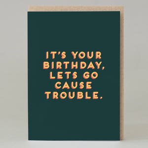 May include: A greeting card with a dark teal background and the text "IT'S YOUR BIRTHDAY, LETS GO CAUSE TROUBLE." in orange and yellow letters. The card has a light brown border.