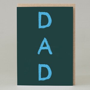 Puede incluir: Una tarjeta de felicitación con la palabra "DAD" en letras azul claro, estilo tiza, sobre un fondo verde oscuro. La tarjeta está colocada sobre una cartulina marrón claro natural.