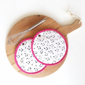 May include: Two white and black patterned pot holders with a pink trim. The pot holders are on a wooden cutting board with a whisk.