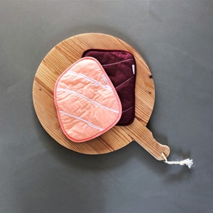 May include: A wooden chopping board with a handle, featuring two fabric pot holders. One pot holder is salmon-coloured with white stripes, and the other is burgundy.
