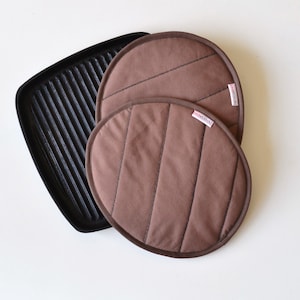 Puede incluir: Dos agarraderas de olla acolchadas de color marrón con el texto "redstitch" en ellas. Las agarraderas están encima de una sartén de hierro fundido negra.