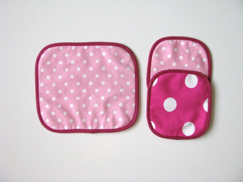 May include: Three pink and white polka dot pot holders. The two smaller pot holders are stacked on top of each other. The larger pot holder is rectangular and the two smaller pot holders are round.